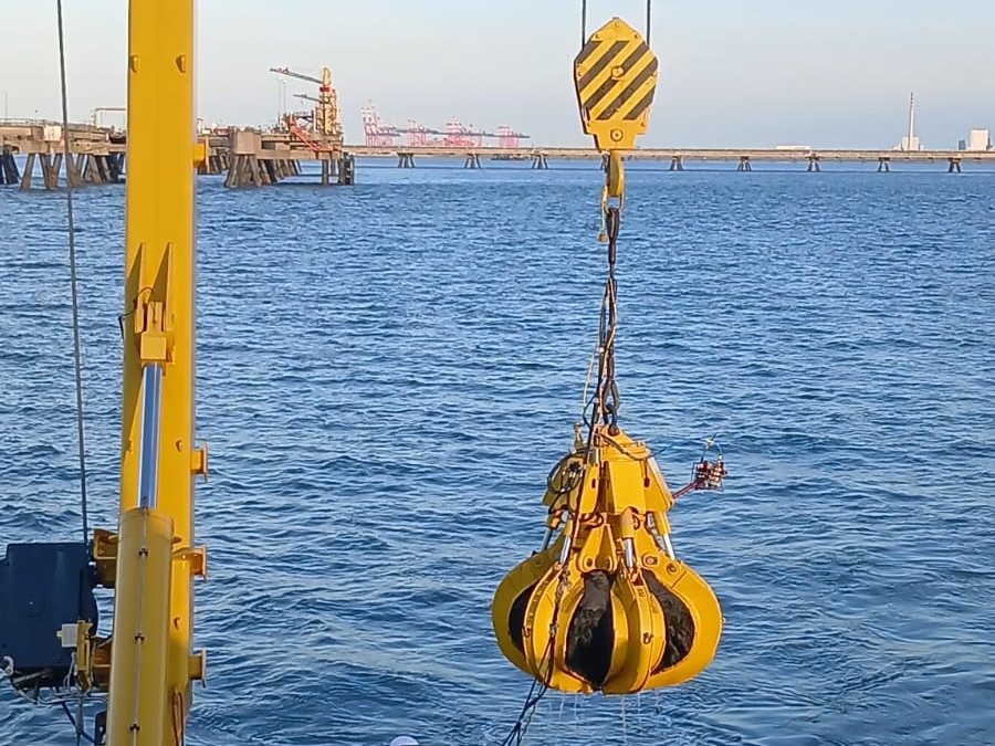 A hydraulic grab lowered from the deck crane, harbour jetties behind